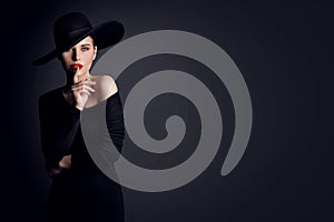 Fashion Woman Model in Hat showing Shh Sign of Silent Gesture putting finger in Red Lips. Elegant Lady in Black Dress over Gray