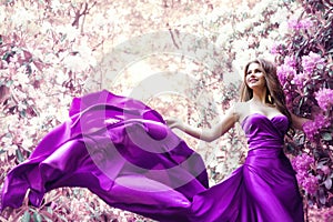 Fashion Woman Pink Dress in Spring Flower Garden. Beauty Model Girl in Purple Silk Gown Fluttering on Wind. Floral Pink Fantasy