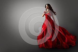 Fashion Woman in Red Dress. Beauty Model dancing in Long Evening Gown fluttering on Wind Black Hair flying in Air over Gray