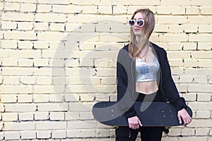 Fashion woman with a skateboard stand on brickwall background. Girl in sunglasses with longboard