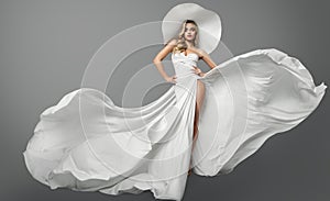 Fashion Woman in White Flying Dress and Summer Hat over Gray Studio Background. Elegant Bride in Wedding Gown. Beautiful Girl in