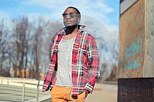 Fashion young african man listens to music in earphones outdoors wearing a plaid red shirt and sunglasses street