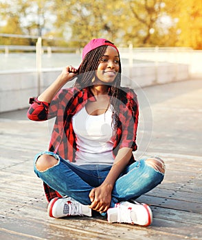Fashion young african woman having fun in the city park, wearing a red checkered shirt cap sitting