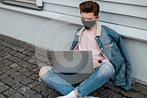Fashion young freelancer man in black medical mask in stylish denim casual jeans clothes sits with laptop and works remotely on