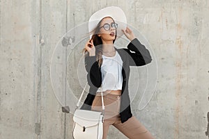 Fashion young girl with beauty sexy face wih leather bag in glasses in stylish black blazer straightens summer beautiful hat near