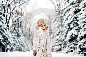 Fashion young smiling blonde woman in winter. Standing among snowy trees in winter forest. Wearing beige coat, white scarf in boho