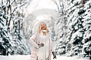 Fashion young smiling blonde woman in winter. Standing among snowy trees in winter forest. Wearing beige coat, white scarf in boho