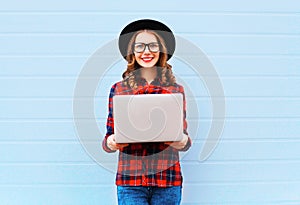 Fashion young smiling woman holding laptop computer in city, wearing black hat red checkered shirt over blue background