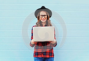 Fashion young smiling woman holding laptop computer in city, wearing a black hat, red checkered shirt over blue background