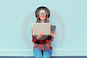 Fashion young smiling woman holding laptop computer with coffee cup resting outdoors in city, wearing black hat red checkered
