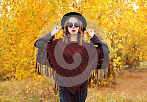 Fashion young woman in autumn day wearing a black hat sunglasses and knitted poncho over yellow leaves