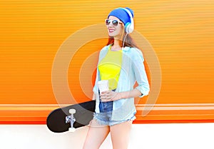 Fashion young woman with skateboard and coffee of cup is listens to music over colorful orange