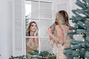 Fashion young woman in stylish gold dress looks at reflection in the mirror by the Christmas trees