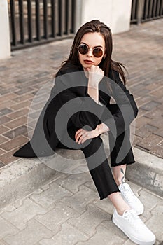 Fashion young woman in trendy pants in stylish youth blazer sitting on stone tiles in city. Fashionable new collection of women`s