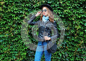 Fashion young woman wearing a black rock jacket, handbag posing over green leaves wall