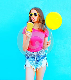 Fashion young woman wearing a t-shirt, denim shorts drinks fruit juice from cup with yellow air balloon over colorful blue