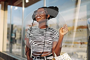 Fashionable African American woman in sunglasses, with shopping bags looking at a shop window in the city