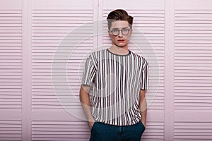 Fashionable American young hipster man in stylish glasses with trendy hairstyle in a vintage striped t-shirt posing near a wooden