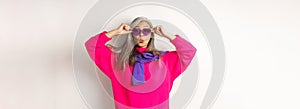 Fashionable asian senior woman in sunglasses and stylish pink sweater pucker lips, raising eyebrows and looking