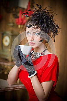 Fashionable attractive young woman in red dress drinking coffee in restaurant. Beautiful brunette in elegant vintage scenery