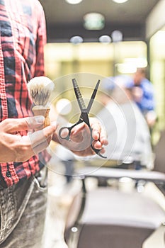 Fashionable barber holding scissors and shaving brush in barbershop. Space for copy