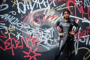 Fashionable beautiful african american woman posing in black leather jacket and pants at street against graffiti wall