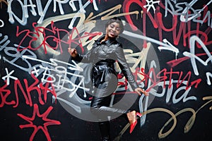 Fashionable beautiful african american woman posing in black leather jacket and pants at street against graffiti wall