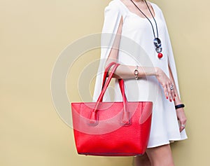 Fashionable beautiful big red handbag on the arm of the girl in a fashionable white dress, posing near the wall on a