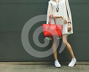 Fashionable beautiful big red handbag on the arm of the girl in a fashionable white dress, posing near the wall on a