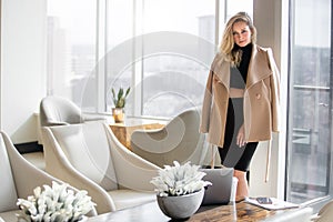 Fashionable beautiful classy, high class woman in coat in hotel lobby with windows showing city buildings in background
