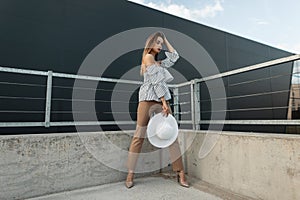 Fashionable beautiful stylish girl in trendy summer clothes holding a hat in the city near the modern building