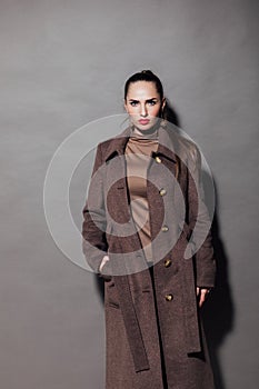 a fashionable and beautiful woman in a brown cape stands on a gray background