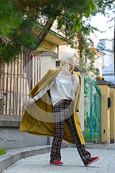 Fashionable beautiful young woman with blond hair in a stylish long coat, checkered pants, red shoes and glasses poses