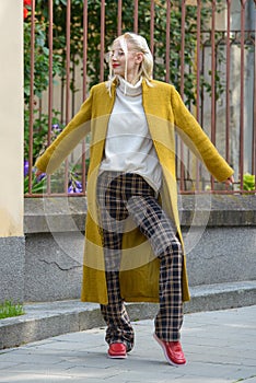 Fashionable beautiful young woman with blond hair in a stylish long coat, checkered pants, red shoes and glasses poses
