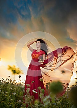 Fashionable beautiful young woman in long red dress posing outdoor with cloudy dramatic sky in background. Attractive brunette