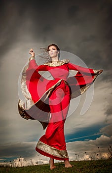 Fashionable beautiful young woman in red long dress posing outdoor with cloudy dramatic sky in background