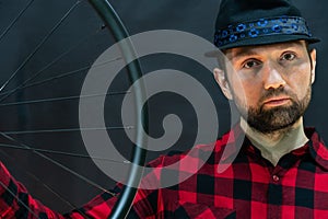 A fashionable bicycle mechanic in a red shirt and a black hat holds a wheel in his hand. Assembling wheels from a carbon rim using