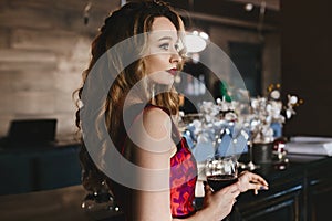 Fashionable blonde model girl with red lips and with curly hairstyle, in the red-pink dress stands at the bar with