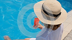 fashionable blonde woman in straw hat relaxes and drinks orange cocktail near the swimming pool on a sunny summer day