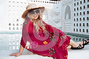 Fashionable blonde woman wearing a colorful swim coverup, sunglasses and beach hat relaxes by a resort swimming pool