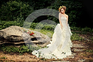 Fashionable bride in a lush white dress is standing by a large stone.