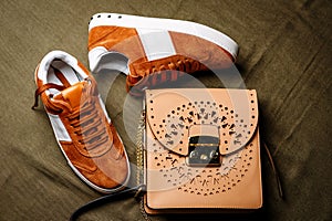 Brown suede sneakers with white accents on a white sole and a brown leather bag with a golden lock on a green woven background