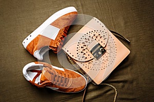 Brown suede sneakers with white accents on a white sole and a brown leather bag with a golden lock on a green woven background