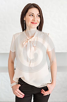 Fashionable brunette girl in a light blouse stands against a white wall, posing and looking at the camera