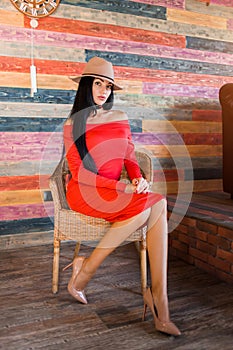 Fashionable brunette woman with long hair and red dress sitting on a chair at home and looking away