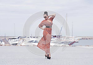 Fashionable brunette woman wearing long dress in sunglasses posing near sea, pier with yachts