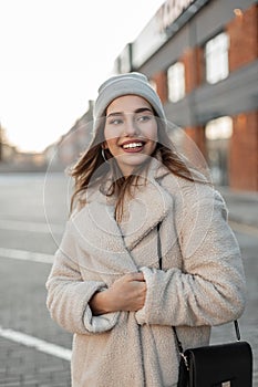 Fashionable cheerful young woman in a vintage hat in a trendy coat with a fashion black leather handbag posing and smiling in a