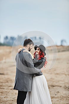 A fashionable couple of newlyweds posing outdoors. Stylish bearded man and his red-haired girlfriend in a wedding dress