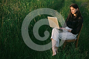 Fashionable elegant girl working on laptop and sitting on rustic chair  in summer field at sunset. Young business woman working