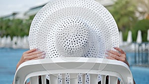 Fashionable female sunbathing in white elegance hat relaxing on deck chair near swimming pool